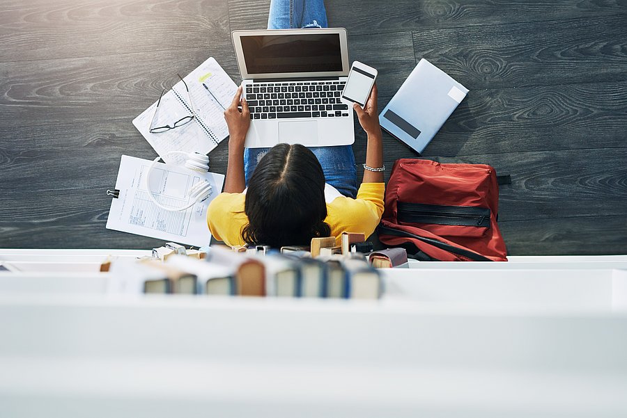 Eine Frau sitzt auf dem Boden mit einem Laptop auf dem Schoß. Um sie herum liegen viele Unterlagen aus Papier und ein Ruchsack