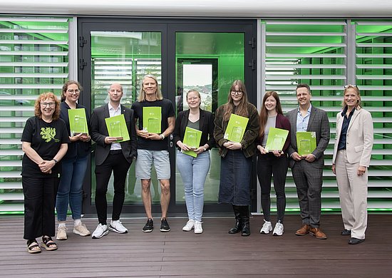 Dr. Katharina Bosbach und weitere Lehrende, die eine Auszeichnung erhalten haben stehen gemeinsam in einem Halbkreis. Alle halten ihre Urkunden und Preisen und lächeln in die Kamera