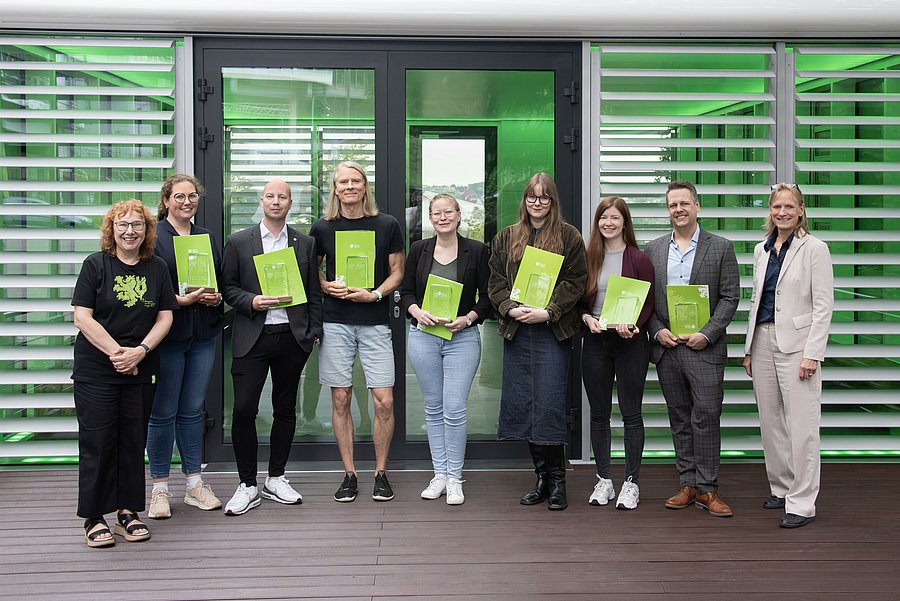 Dr. Katharina Bosbach und weitere Lehrende, die eine Auszeichnung erhalten haben stehen gemeinsam in einem Halbkreis. Alle halten ihre Urkunden und Preisen und lächeln in die Kamera