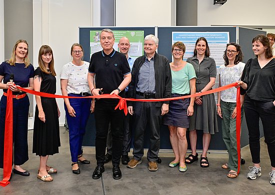 Alle Professorinnen und Professoren des Instituts stehen hinter einem roten Band. Professor Schulze hält eine Schere in der Hand, um das rote Band aufzuschneiden zur Eröffnung der neuen Räumlichkeiten