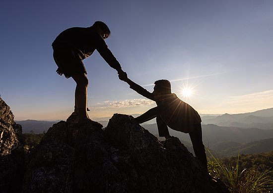 Zwei Personen klettern einen Felsen hoch. Eine Person hilft dabei der anderen Person und zieht sie hoch