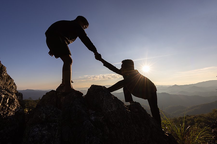 Zwei Personen klettern einen Felsen hoch. Eine Person hilft dabei der anderen Person und zieht sie hoch