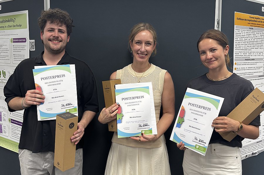 Luka Frischko, Ann-Kathrin Sowa und Sarah Lirien Wünsche stehen vor einer Posterwand. Alle halten eine Urkunde sowie einen Preis in den Händen. 
