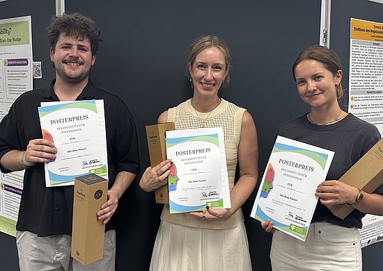 Luka Frischko, Ann-Kathrin Sowa und Sarah Lirien Wünsche stehen vor einer Posterwand. Alle halten eine Urkunde sowie einen Preis in den Händen. 