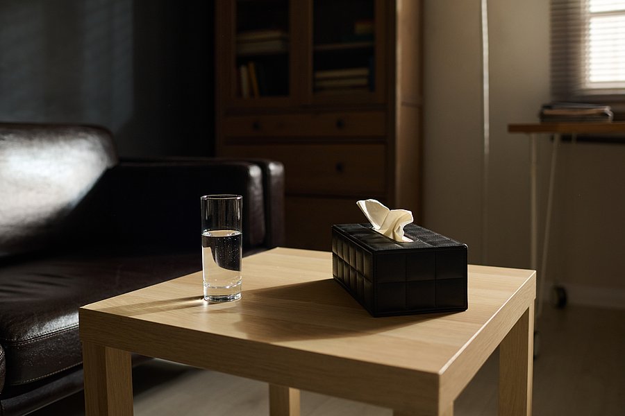 Ein Raum mit einem Couchtisch um Fokus. Darauf steht ein Glas mit Wasser und eine Box mit Taschentücher. Im Hintergrund steht ein Sofa