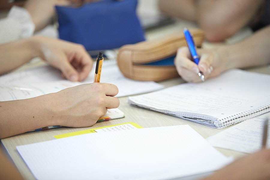 Auf einem Tisch sind mehere Notizblöcke zu sehen. Studierende notieren sich darauf etwas, man sieht jedoch nur die Arme und die Stifte