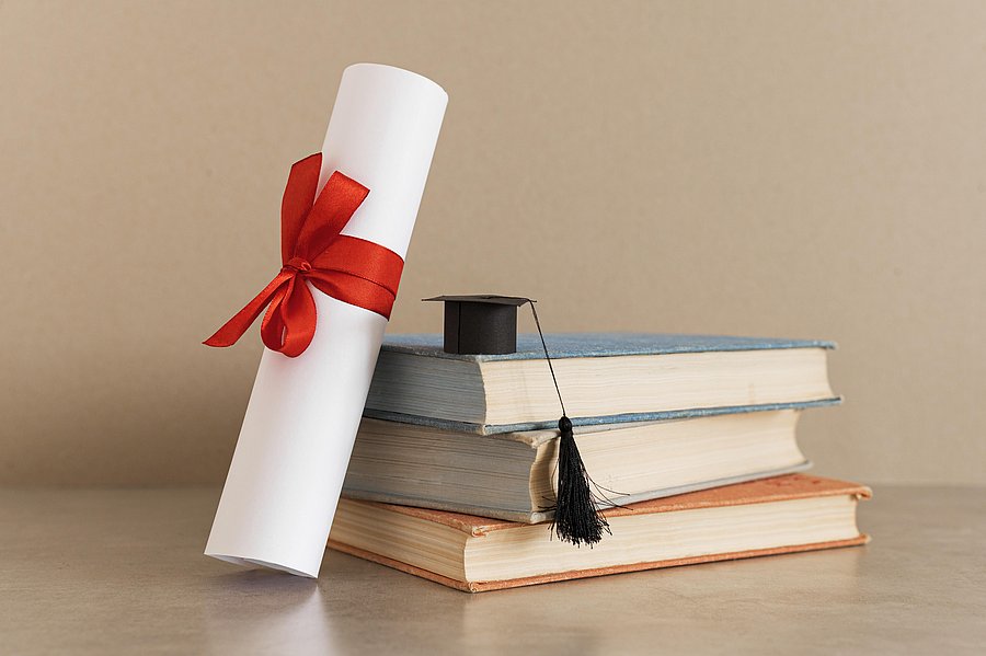 Eine Urkunde, eingerollt mit einer roten Schleife, lehnt gehgen einen Stapel mit drei Büchern
