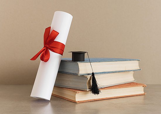 Eine Urkunde, eingerollt mit einer roten Schleife, lehnt gehgen einen Stapel mit drei Büchern