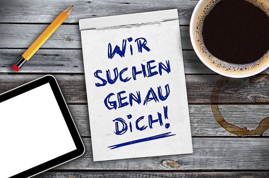 Auf einem Tisch liegt ein Notizbuch; auf diesem steht: "Wir suchen genau dich". Daneben liegen ein Stift und ein Tablet. Eine Tasse Kaffee steht daneben