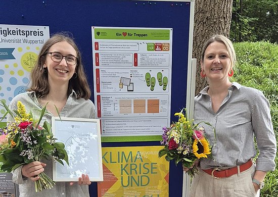 Dana Braß und Prof. Dr. Theda Radtke stehen vor einem Poster, welches ihr Projekt "Ein Herz für Treppen" beschreibt. Beide halten einen Blumentstrauß in der Hand und lächeln in die Kamera.