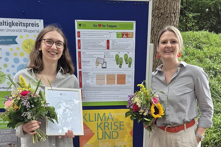 Dana Braß und Prof. Dr. Theda Radtke stehen vor einem Poster, welches ihr Projekt "Ein Herz für Treppen" beschreibt. Beide halten einen Blumentstrauß in der Hand und lächeln in die Kamera.